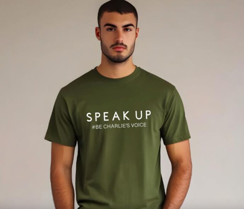 Puede incluir: Camiseta verde oliva con las palabras "SPEAK UP" y el hashtag "#BE CHARLIE'S VOICE" impresos en blanco. La camiseta tiene mangas cortas y cuello redondo.