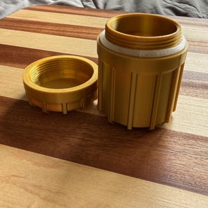 May include: A gold-colored cylindrical container with a matching screw-on lid. The container features vertical ridges along its exterior and a white threaded rim at the top. The lid is detached and placed beside the open container on a flat surface.