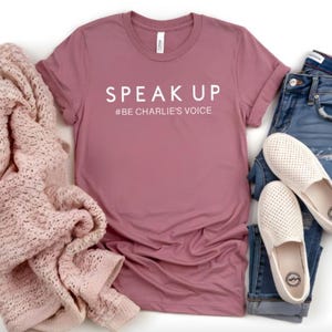Puede incluir: Una camiseta color malva con las palabras "SPEAK UP" y el hashtag "#BE CHARLIE'S VOICE" en blanco. La camiseta se combina con vaqueros azules, zapatos blancos sin cordones y una manta de punto rosa. Un atuendo informal.