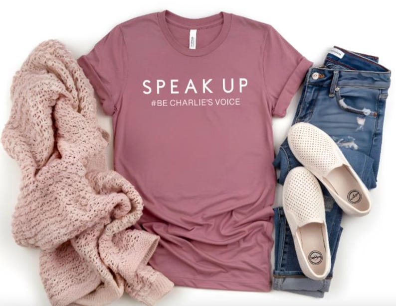 Puede incluir: Una camiseta color malva con las palabras "SPEAK UP" y el hashtag "#BE CHARLIE'S VOICE" en blanco. La camiseta se combina con vaqueros azules, zapatos blancos sin cordones y una manta de punto rosa. Un atuendo informal.