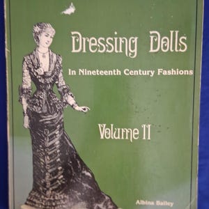 May include: A book cover with a green background featuring the title "Dressing Dolls" and the subtitle "In Nineteenth Century Fashions." A detailed illustration of a woman in a period dress is on the cover. The book is labeled "Volume II" and the author is Albina Bailey.
