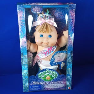 May include: A Cabbage Patch Kids Millennium Celebration doll in its original box. The doll has blonde hair, blue eyes, and a pink sash that says "Happy 2000". The box features a fireworks design and the words "Collector Edition".