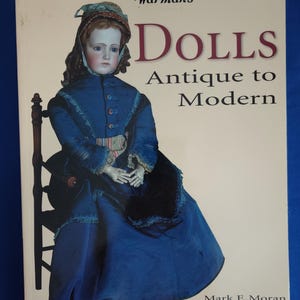 May include: A book cover featuring a vintage doll in a blue dress and bonnet. The title reads "Warman's Dolls Antique to Modern" with the subtitle "Identification and Price Guide" by Mark F. Moran. The cover has a cream background.