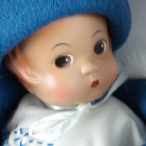 Könnte beinhalten: Eine Vintage-Puppe mit blauem Hut und passendem Outfit. Die Puppe hat einen hellen Teint, rosige Wangen und rote Lippen. Das Outfit umfasst einen weißen Kragen und ein blau-weißes Karomuster.