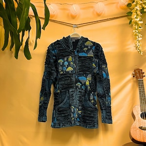 May include: A dark blue denim jacket with a zip fastening and a mushroom print. The jacket has a hood and is hanging on a hanger.