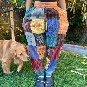 May include: Multicolored patchwork pants with a mushroom print. The pants have an elastic waistband and a drawstring. The pants are worn by a person standing on a grassy lawn.