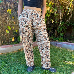 May include: Beige pants with a mushroom and peace sign pattern. The pants have an elastic waistband and a relaxed fit.
