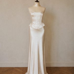 May include: A floor-length, cream-colored satin gown featuring a strapless corset-style bodice with vertical seams, a flared peplum waist detail, and a smooth, flowing skirt that extends to the floor, displayed on a dress form mannequin.