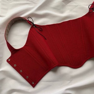 Könnte beinhalten: Ein rotes Korsett-Top mit strukturiertem Design. Das Oberteil hat einen geschwungenen Ausschnitt, Ösen und schwarze Bindebänder. Das Innenfutter ist in einem neutralen Beigeton gehalten. Das Korsett ist aus einem strukturierten Stoff gefertigt.