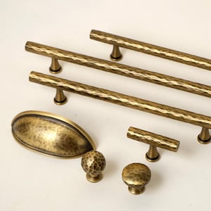 May include: A collection of antique brass cabinet hardware, including bar pulls, knobs, and a cup pull. The hardware has a hammered texture and a warm, aged finish. The arrangement showcases various sizes and styles for kitchen or furniture projects.