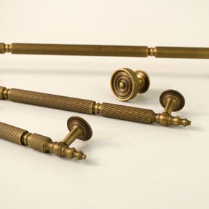 Pode incluir: Close-up de várias peças de ferragens de armário em latão antigo. A coleção inclui puxadores longos, um botão redondo e puxadores menores com um design decorativo canelado. As ferragens têm um acabamento quente e envelhecido.