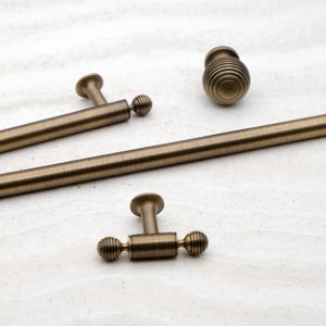 May include: Close-up of antique brass cabinet hardware, including a knob and several pulls. The pulls have a T-shaped base with a ribbed, spherical end. The knob has a ribbed, spherical design. The hardware is displayed on a light-colored surface.
