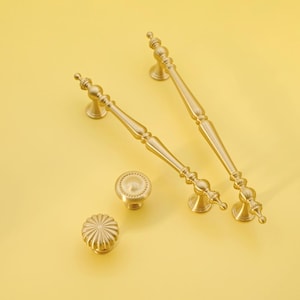 Peut inclure: Ensemble de quincaillerie de placard dorée, comprenant deux longues poignées et deux boutons. Les poignées ont un design décoratif et les boutons présentent une surface ronde et texturée. L'ensemble est présenté sur un fond jaune.