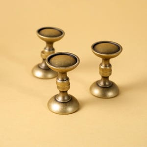 May include: Three antique brass-colored candle holders are arranged on a light yellow surface. Each holder has a round base, a central stem, and a circular top. The tops have a slightly raised, textured edge. The overall design is vintage.