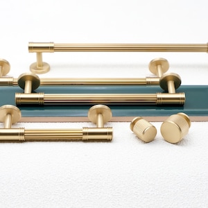 May include: A selection of gold-coloured cabinet hardware, including bar pulls and knobs. The pulls have a ribbed design and are mounted on a green and white surface. The knobs are cylindrical with a textured pattern. The hardware is for furniture.