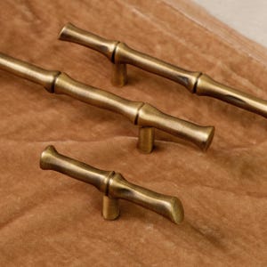 Puede incluir: Cuatro tiradores de armario de latón antiguo con diseño de bambú. Los tiradores están dispuestos en diagonal, mostrando sus superficies detalladas y texturizadas, de color marrón dorado cálido. El herraje está sobre un fondo marrón.