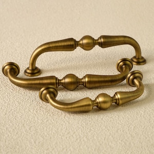 May include: Three antique-style bronze cabinet pulls. Each pull features a curved handle with decorative ball and cylindrical elements. The hardware is designed for easy installation and adds a vintage touch to furniture.