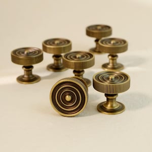 Op de afbeelding: Een verzameling van zeven antieke messing kastknoppen. Elke knop heeft een cirkelvormig ontwerp met concentrische ringen en een geribbelde rand, gemonteerd op een korte voet. De knoppen hebben een warme, verouderde messingkleur.