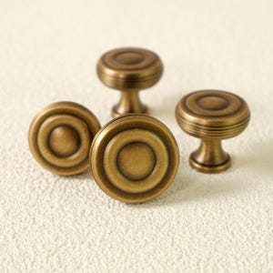 Op de afbeelding: Vier antieke messing kastknoppen met een cirkelvormig ontwerp. De knoppen hebben een warme, verouderde afwerking en zijn gerangschikt op een getextureerd, gebroken wit oppervlak. Een klassieke hardware keuze.