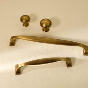 Może przedstawiać: Zestaw antycznych mosiężnych okuć do szafek, w tym dwa gałki i dwa uchwyty. Gałki są okrągłe z lekko wypukłym wierzchem. Uchwyty mają zakrzywiony kształt. Okucia mają ciepłe, postarzane wykończenie.