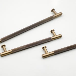 Puede incluir: Cuatro tiradores de gabinete de color bronce con un diseño acanalado. Cada tirador tiene un mango cilíndrico y una base en forma de T. El herraje se encuentra sobre un fondo blanco, mostrando los detalles de los tiradores decorativos.