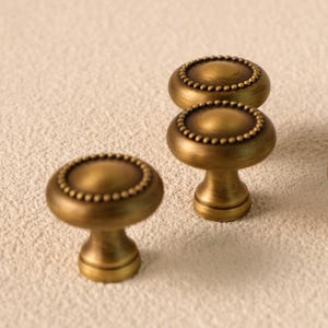 Könnte beinhalten: Nahaufnahme von drei antiken Messing-Schrankknöpfen. Jeder Knopf hat eine abgerundete Oberseite mit einem Perlenrand und einen kurzen, zylindrischen Sockel. Die Knöpfe haben eine warme, goldene Farbe, die auf eine gealterte Oberfläche hindeutet.