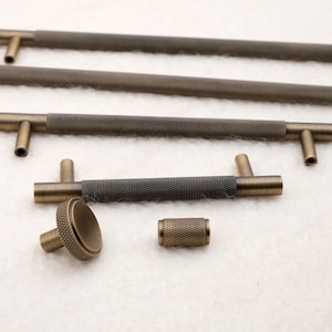 Peut inclure: Ensemble de quincaillerie de meubles de couleur bronze, comprenant plusieurs barres de tirage et un bouton. Les poignées ont une prise centrale texturée et mesurent environ 15 cm de long. Le bouton et la petite pièce cylindrique ont également une surface texturée.