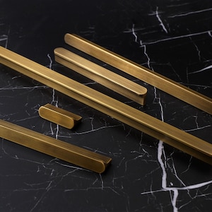 Può includere: Diverse maniglie e pomelli per mobili in ottone di varie lunghezze sono disposti su una superficie di marmo nero. L'hardware ha una forma a barra rettangolare con estremità arrotondate, suggerendo un'estetica di design moderno. La calda finitura metallica contrasta con lo sfondo scuro.