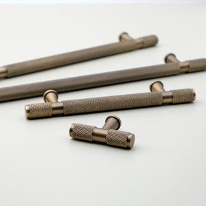 May include: Close-up of several antique brass cabinet pulls and knobs. The pulls are long and cylindrical with a textured surface. The knobs are T-shaped with a similar textured design. The hardware is arranged on a light background.