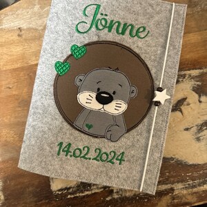 Könnte beinhalten: Eine graue Filzhülle mit dem Namen "Jönne" in grüner Schrift gestickt. Ein brauner Kreis zeigt einen grauen Cartoon-Otter mit herzförmiger Nase und grünen Herzakzenten. Das Datum "14.02.2024" ist grün gestickt.