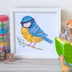 May include: A framed artwork featuring a bluebird crafted from various blue and yellow buttons. The bird is perched on a brown branch with green leaves, set against a white background. The artwork is in a white frame, with toy animals in the background.