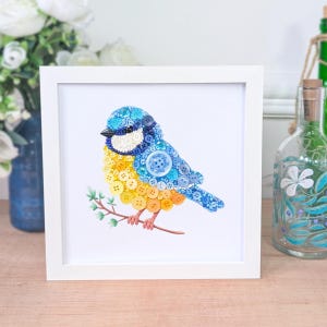 May include: A square white frame containing a mosaic artwork of a bird perched on a branch. The bird is constructed from numerous small buttons in shades of blue, yellow, and white, featuring a black beak and brown feet on a brown branch.