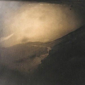 May include: A black and white photograph of a landscape with a winding path leading up a hill. The sky is cloudy and the hills are covered in a light mist.