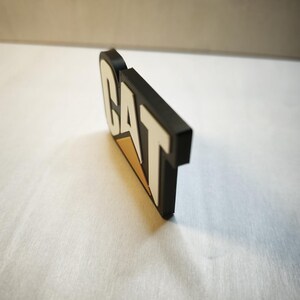 Pu&ograve; includere: Un logo CAT in bianco e nero con un accento triangolare giallo. Il logo &egrave; un design tridimensionale, con le lettere in rilievo e i bordi arrotondati. Il logo &egrave; posto su uno sfondo bianco.