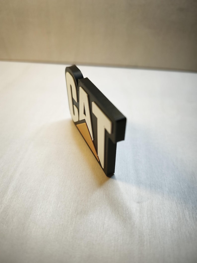 Pu&ograve; includere: Un logo CAT in bianco e nero con un accento triangolare giallo. Il logo &egrave; un design tridimensionale, con le lettere in rilievo e i bordi arrotondati. Il logo &egrave; posto su uno sfondo bianco.