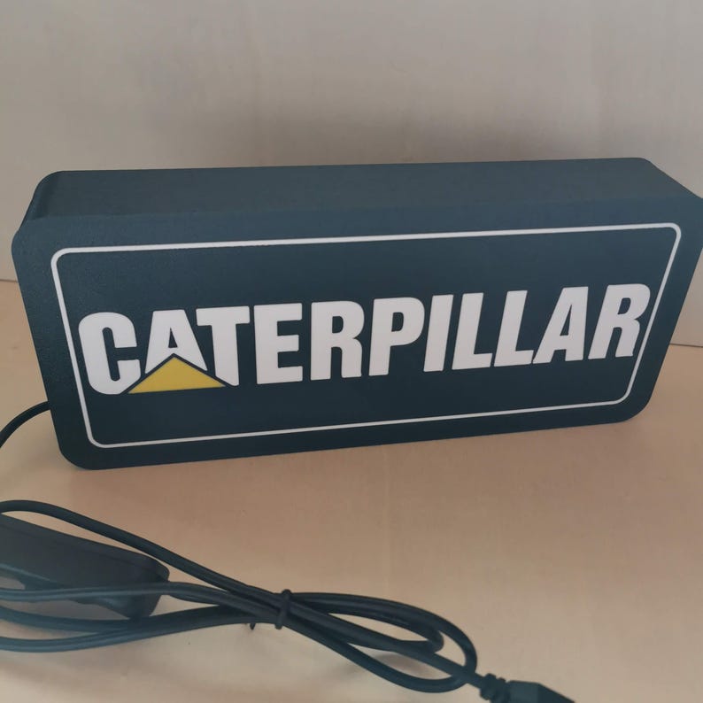 Pu&ograve; includere: Un cartello rettangolare blu scuro con la scritta "CATERPILLAR" in bianco e un logo giallo e bianco. Il cartello ha un bordo bianco e un cavo di alimentazione nero con un interruttore. Il cartello &egrave; illuminato.