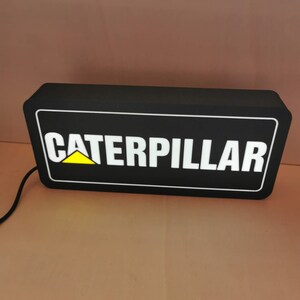 Pu&ograve; includere: Cartello rettangolare nero con la scritta "CATERPILLAR" in bianco, bordata di bianco, con un logo a triangolo giallo. Il cartello &egrave; illuminato e ha un cavo di alimentazione.