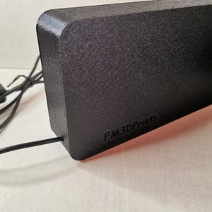 Pu&ograve; includere: Un oggetto rettangolare nero con angoli arrotondati, possibilmente stampato in 3D. Il testo "FM3DPrints" &egrave; visibile sul lato. Un cavo nero esce dall'oggetto, suggerendo che sia elettronico.