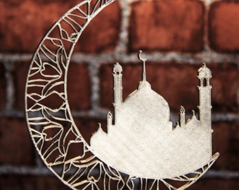 May include: A silver metal ornament shaped like a crescent moon with a detailed filigree design. The moon is holding a silhouette of a mosque with multiple minarets.