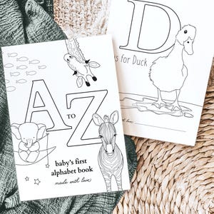 Può includere: Due libri da colorare aperti con disegni a tratto nero. Un libro mostra le lettere dalla A alla Z con illustrazioni di animali. L'altro libro mostra la lettera D con un'anatra. Il testo "baby's first alphabet book" è visibile.