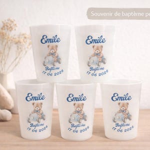 Peut inclure: Gobelets en plastique blancs avec un motif d'ours en peluche et le nom "Emile" en bleu. Les gobelets portent également l'inscription "Baptême 17 06 2026". Un lapin en peluche et des fleurs séchées sont en arrière-plan.