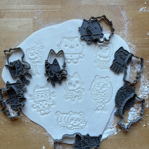 Peut inclure: Des découpoirs à biscuits en plastique gris en forme de chats, disposés autour de formes de chats découpées dans de la pâte blanche. Les découpoirs présentent divers motifs de chats, en position debout, assise et couchée. De la farine est saupoudrée sur la surface en bois.