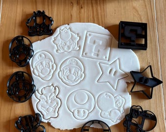 May include: A collection of black plastic cookie cutters in various shapes, including Mario, Princess Peach, and a question mark block. The cutters are arranged around a piece of rolled-out dough, with a wooden rolling pin in the background. The theme is related to the Super Mario video game series.