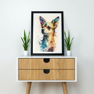 May include: A watercolor painting of a chihuahua dog with colorful splashes of paint in the background. The painting is framed in a black frame and is displayed on a wooden dresser with two drawers.