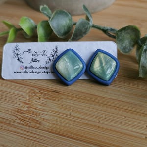 May include: A pair of square stud earrings with a matte blue border and a shimmering, pearlescent light green center. The earrings are attached to a white display card featuring the text Nilico, @nilico_design, and www.nilicodesign.etsy.com.