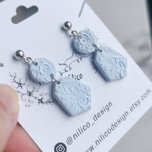 May include: Pale blue earrings with a floral design and silver-tone findings. Each earring features a circular top and a hexagonal bottom, linked by a small chain. The earrings are presented on a white card with the text "nilico_design" and a website address.