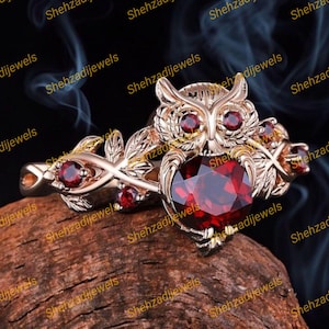 May include: A rose gold owl ring featuring a large, heart-shaped, red gemstone. The owl's eyes are small red gemstones. The ring has a detailed, nature-inspired design with leaves and additional red gemstones.