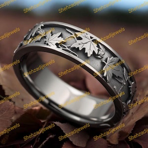 May include: A silver-toned ring with a detailed leaf design. The ring features a band with raised leaf patterns and gold-colored accents. The ring is set against a backdrop of autumn leaves, creating a nature-inspired aesthetic.