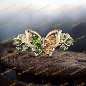 May include: A gold-toned ring featuring a butterfly design with green and yellow gemstones. The butterfly's wings are detailed with openwork, and the ring is adorned with additional green gemstones on the sides. The text "Shehzadijewels" is repeated across the image.