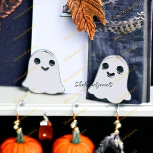 Puede incluir: Dos sujetalibros de plata en forma de fantasma con ojos negros y una boca sonriente. Los sujetalibros están en un estante con dos pendientes en forma de calabaza naranja colgando debajo.